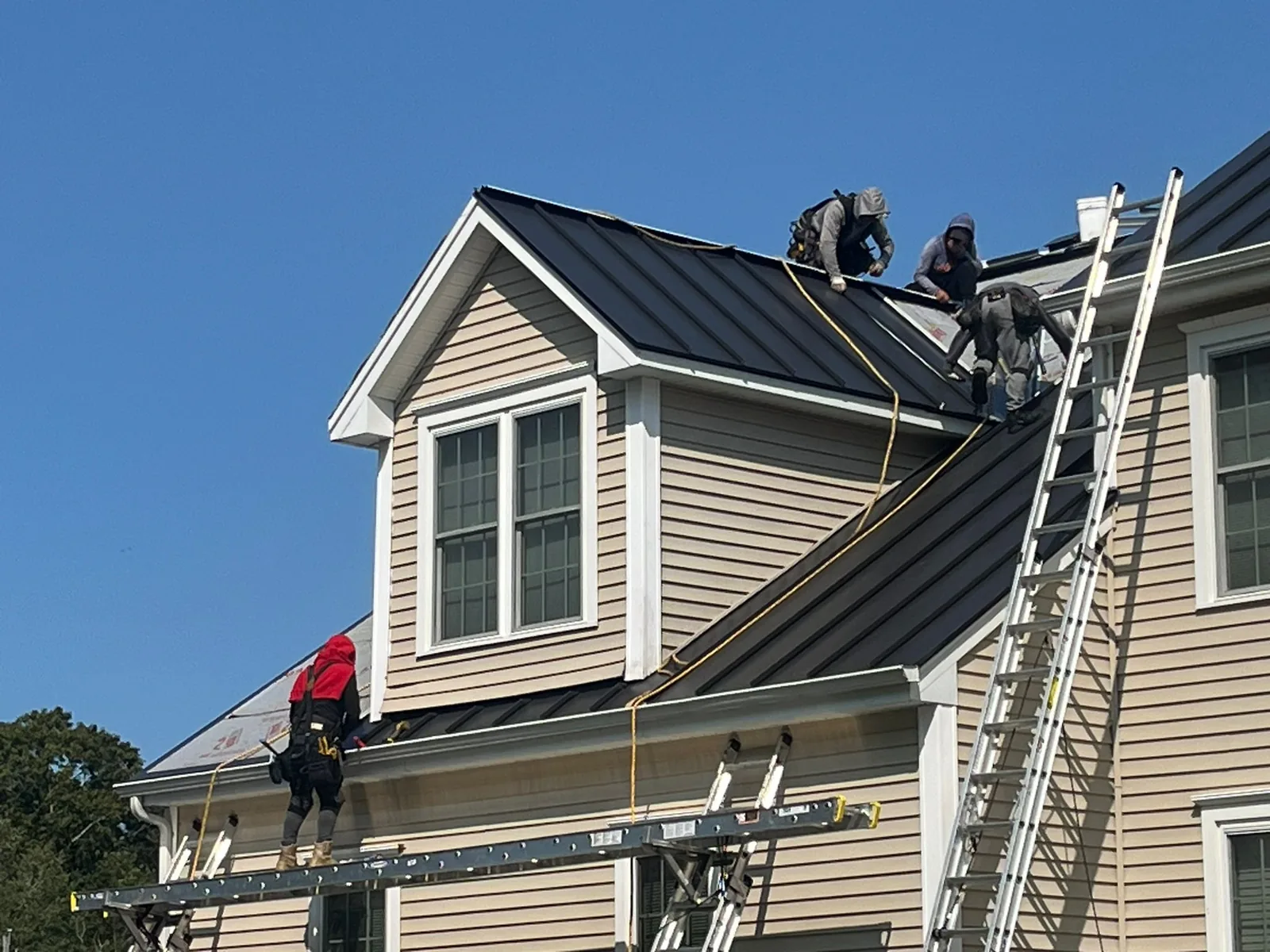 Roofing crew in safety harnesses installing black standing seam metal roofing panels on a two story house